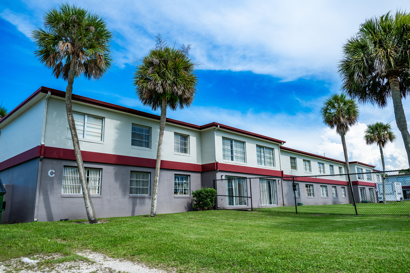 The back side of a two story building with sliding glass doors on the ground floor. The building is surrounded by green grass and palm trees.